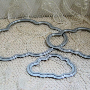 May include: Three metal cloud-shaped die-cut stencils. The largest stencil is silver and has a wavy edge. The two smaller stencils are nested inside the larger stencil and have a more rounded edge.