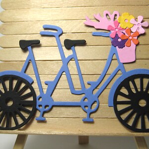 May include: A blue tandem bicycle with black wheels and a pink basket filled with colorful paper flowers. The bicycle is mounted on a wooden easel.