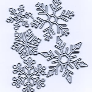 May include: A collection of six silver-colored snowflake die cuts in various sizes. Each snowflake features intricate, openwork designs with detailed edges. The snowflakes are arranged on a white background, ideal for winter-themed crafts and decorations.