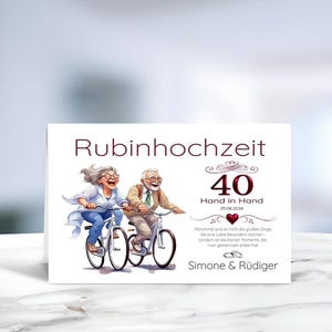 Könnte beinhalten: Eine weiße Grußkarte mit einer Illustration eines älteren Paares auf Fahrrädern. Der Text lautet Rubinhochzeit, 40, Hand in Hand, 25.08.2026, Simone & Rüdiger sowie ein Zitat. Ein kleines rotes Herz und zwei Ringe sind unter dem Text abgebildet.