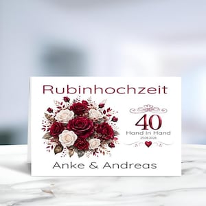 Könnte beinhalten: Eine weiße, rechteckige Grußkarte mit einem Blumenstrauß aus dunkelroten und weißen Rosen. Der Text lautet Rubinhochzeit, 40 Hand in Hand, 25.08.2026 und Anke & Andreas. Das Design enthält dekorative Schnörkel und ein kleines rotes Herzsymbol.