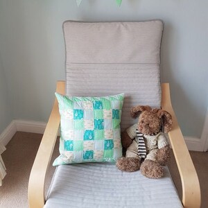 May include: A light grey armchair with a patchwork cushion featuring a green and white pattern. A brown teddy bear with a striped scarf is sitting on the chair. A green and white bunting hangs above the chair.