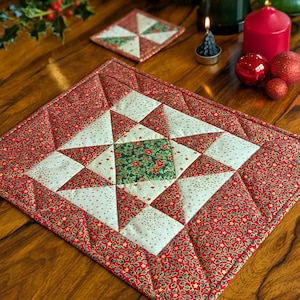 May include: A festive quilted placemat with a star design. The placemat has a red background with a pattern of green leaves and gold stars. The star is made of white and green fabric. A matching coaster is also visible.
