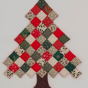 May include: A quilted Christmas tree decoration. The tree is made of red, green, and cream squares with festive patterns. The trunk is brown. A festive holiday decoration.