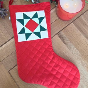 May include: A red quilted Christmas stocking featuring a square patch with a geometric star pattern in white, dark green, and red. The stocking has a fabric loop at the top corner for hanging and a diamond-stitched texture across the entire surface.