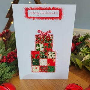 May include: A white greeting card featuring the text Merry Christmas in red font on a fringed red fabric border. Below, a stacked gift box design is created from patchwork fabric squares in red, green, and white with a red and white gingham bow.