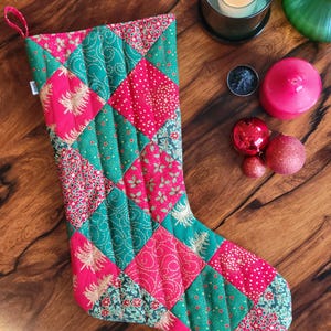 May include: A quilted Christmas stocking featuring a patchwork design of red and green fabric squares. The fabric patterns include gold-coloured Christmas trees, holly leaves, and small stars. A small white tag with the word Sewn is attached.