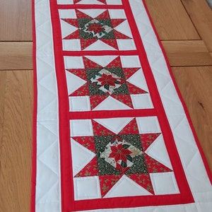 May include: A rectangular Christmas table runner with a repeating star pattern in red and white, featuring poinsettia designs. It has a red border and white quilted sections, creating a festive look. The dimensions are approximately 150 cm.