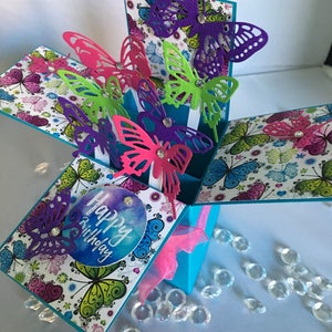 May include: A turquoise blue pop-up card with a happy birthday message and colourful butterflies. The card is decorated with a white background and a butterfly pattern.