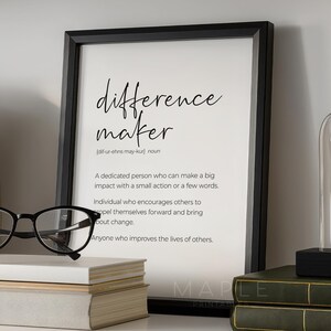 Puede incluir: Una lámina de arte enmarcada en negro que presenta la definición de diccionario del término difference maker en texto negro sobre fondo blanco, expuesta junto a unas gafas negras y dos libros apilados en una estantería.