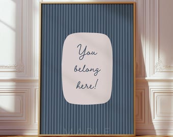 May include: A rectangular wall art print featuring a dark blue background with thin vertical stripes. In the center, a light pink rounded rectangular shape contains the dark blue text "You belong here!" in a cursive, handwritten-style font.