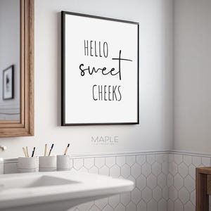 May include: A framed bathroom wall art print with the text "HELLO sweet CHEEKS" in black script. The print is in a black frame and hangs on a white wall above a bathroom sink. The bathroom has a mirror and a small plant.