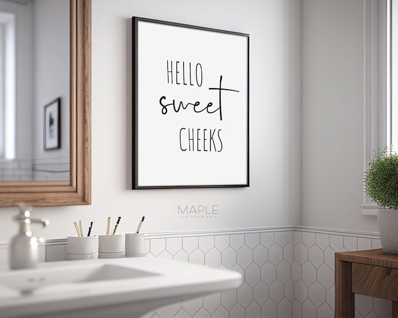 May include: A framed bathroom wall art print with the text "HELLO sweet CHEEKS" in black script. The print is in a black frame and hangs on a white wall above a bathroom sink. The bathroom has a mirror and a small plant.