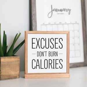 May include: A wooden sign with the text "EXCUSES - DON'T BURN - CALORIES" in black lettering on a white background. The sign is sitting on a white surface next to a potted plant and a framed calendar.