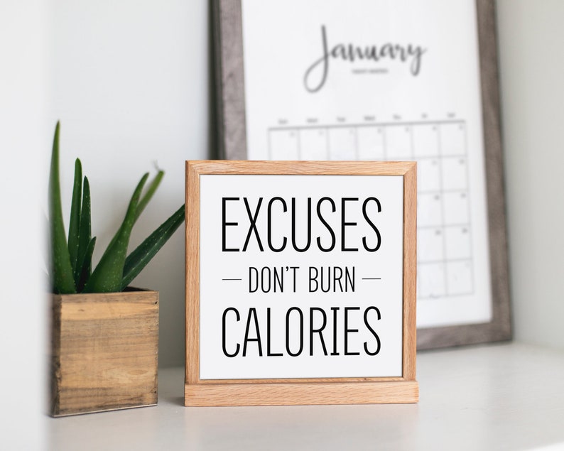 May include: A wooden sign with the text "EXCUSES - DON'T BURN - CALORIES" in black lettering on a white background. The sign is sitting on a white surface next to a potted plant and a framed calendar.
