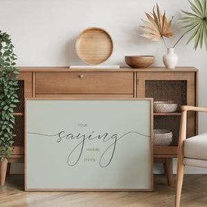 May include: A light green framed print with the text "Your saying where it fits" in a cursive font. The print is hanging on a wall above a wooden cabinet.