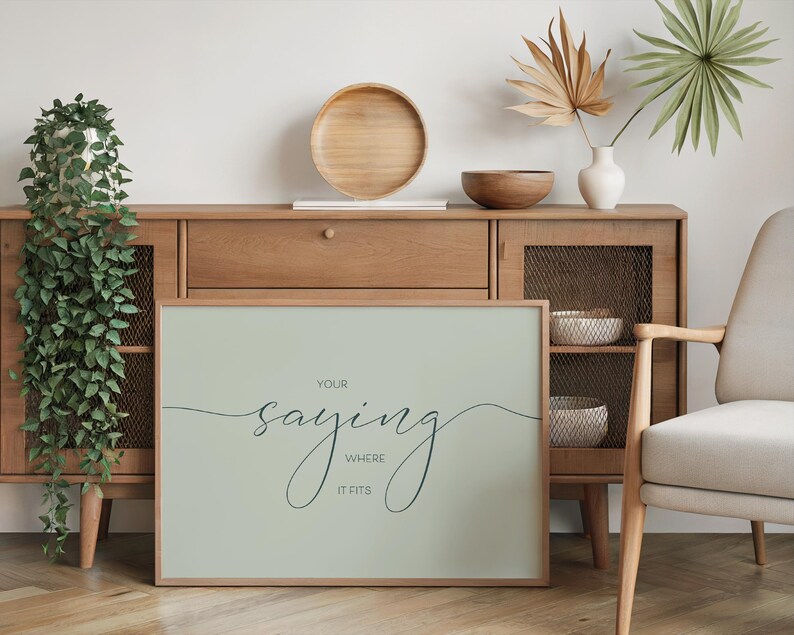 May include: A light green framed print with the text "Your saying where it fits" in a cursive font. The print is hanging on a wall above a wooden cabinet.
