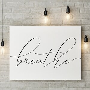 May include: A white canvas print with the word "breathe" written in black cursive script. The print is hanging on a brick wall with several black pendant lights hanging above it.