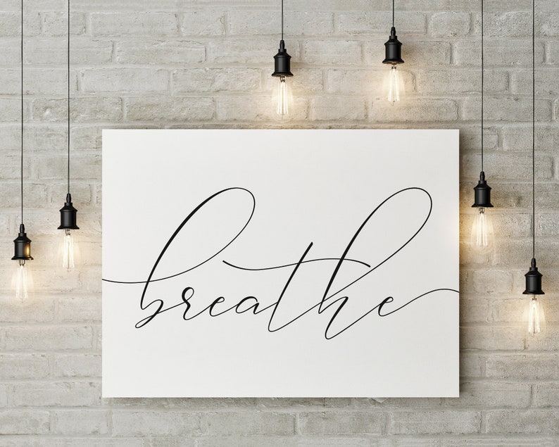 May include: A white canvas print with the word "breathe" written in black cursive script. The print is hanging on a brick wall with several black pendant lights hanging above it.
