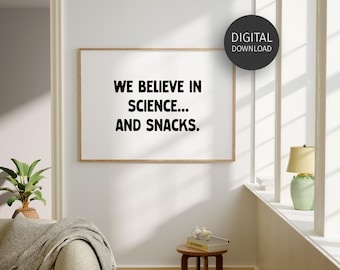 May include: A framed print on a white wall with the text "WE BELIEVE IN SCIENCE... AND SNACKS." The print has a light brown frame. A digital download sticker is in the upper right corner. A small table with books and a lamp are in the room.