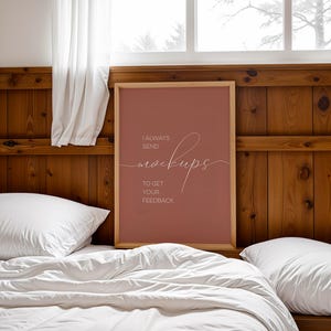 May include: A framed poster with a light brown wood frame and a red background. The poster says "I always send mockups to get your feedback". The poster is hanging on a wooden wall above a white bed with white pillows.