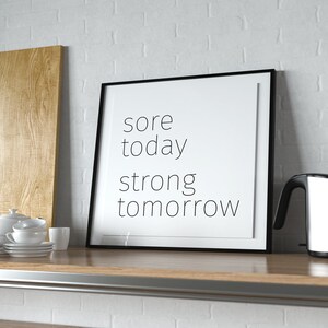 May include: A black framed motivational poster with the text "sore today strong tomorrow" on a white background. The poster is on a wooden shelf with a wooden chopping board, a white teapot, and a silver electric kettle.