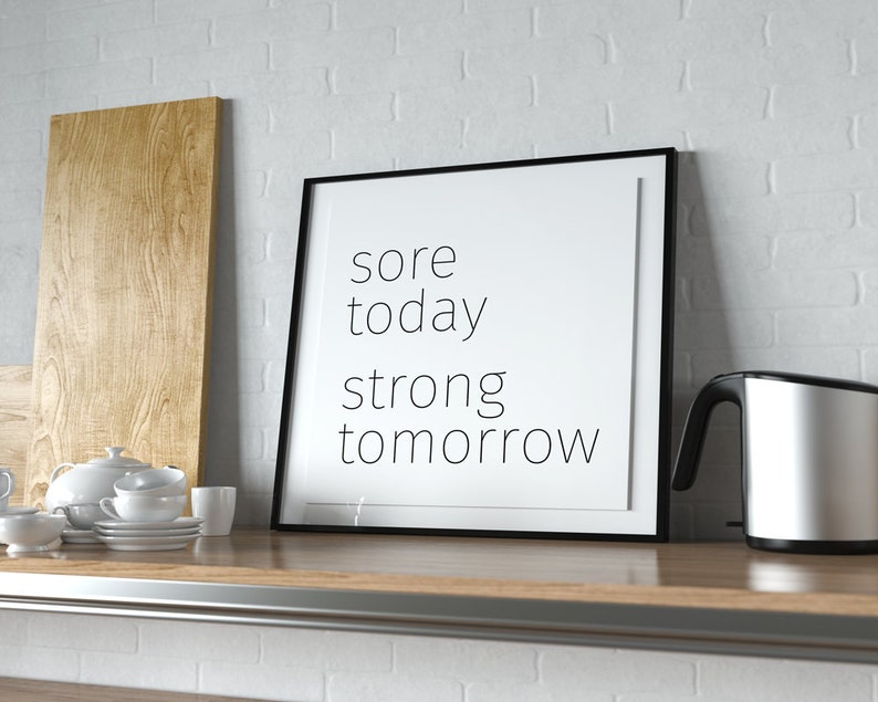 May include: A black framed motivational poster with the text "sore today strong tomorrow" on a white background. The poster is on a wooden shelf with a wooden chopping board, a white teapot, and a silver electric kettle.