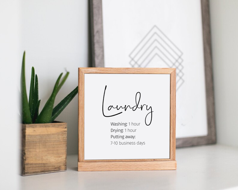 May include: A framed sign with the word "Laundry" in elegant script. Below, the sign lists laundry times: washing 1 hour, drying 1 hour, and putting away in 7-10 business days. A small potted plant and a framed geometric print are in the background.