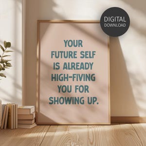 May include: A framed print with the text "YOUR FUTURE SELF IS ALREADY HIGH-FIVING YOU FOR SHOWING UP." The print has a light pink background and teal text. A black circle with "DIGITAL DOWNLOAD" is in the upper right corner. The frame is a light brown color.