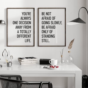 May include: Two black framed motivational posters with white text. The left poster says "You're always one decision away from a totally different life." The right poster says "Be not afraid of going slowly, be afraid only of standing still."