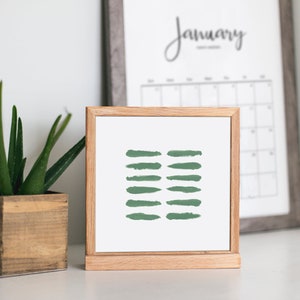 May include: A framed print with a minimalist design featuring a series of green brushstrokes on a white background. The print is displayed on a wooden easel.