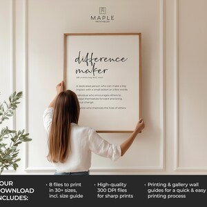 May include: A framed print with the words "difference maker" in a stylish font. The definition of "difference maker" is below the title. The print is being held by a person in a white shirt.