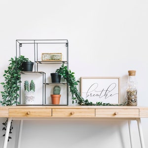 May include: A wooden desk with three drawers and white legs. A black metal shelf unit with plants and a cactus. A framed print with the word "breathe" in black script. A clear glass bottle with a cork top and string lights.