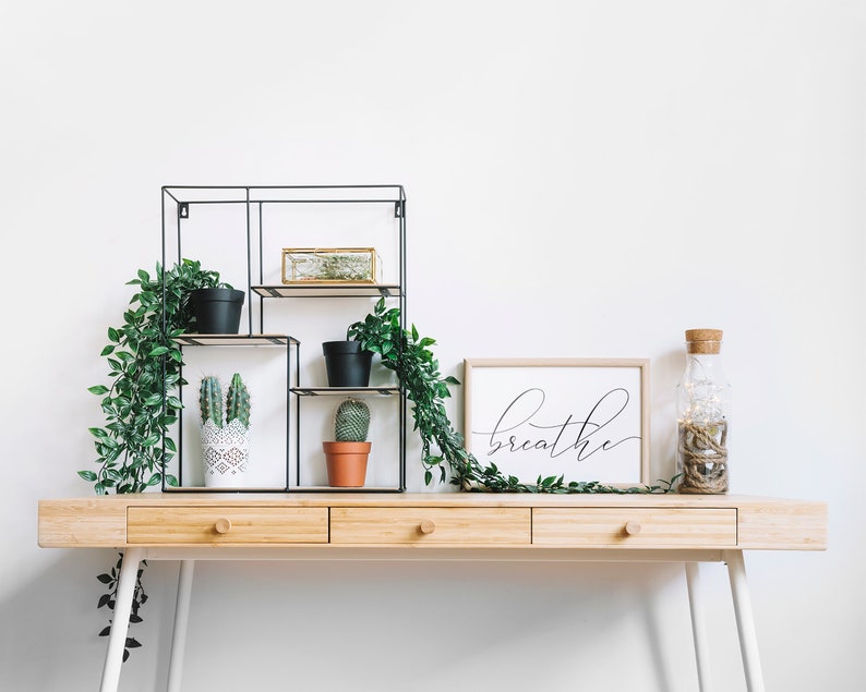 May include: A wooden desk with three drawers and white legs. A black metal shelf unit with plants and a cactus. A framed print with the word "breathe" in black script. A clear glass bottle with a cork top and string lights.