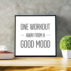 May include: A black framed motivational poster with white text that reads "One workout away from a good mood". The poster is sitting on a wooden table with a white cup and a book.