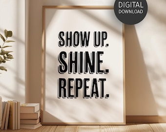 Op de afbeelding: Een ingelijste print met de woorden "SHOW UP. SHINE. REPEAT." in vette zwarte letters. De print heeft een licht houten frame en leunt tegen een muur. Een digitaal download-pictogram staat in de rechterbovenhoek.