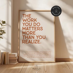 May include: A framed print with the text "THE WORK YOU DO MATTERS MORE THAN YOU REALIZE" in a warm brown colour. The print is displayed on a light-coloured wall, with a stack of books and a plant visible.