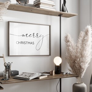 Pu&ograve; includere: Una stampa incorniciata nera con il testo "merry CHRISTMAS" in un carattere corsivo, appesa a una parete bianca sopra una scrivania di legno con una lampada, un vaso di erba della pampa secca e un libro.