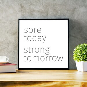 May include: A black framed motivational poster with the text "sore today strong tomorrow" on a white background. The poster is sitting on a wooden table with a white coffee cup and a potted plant.