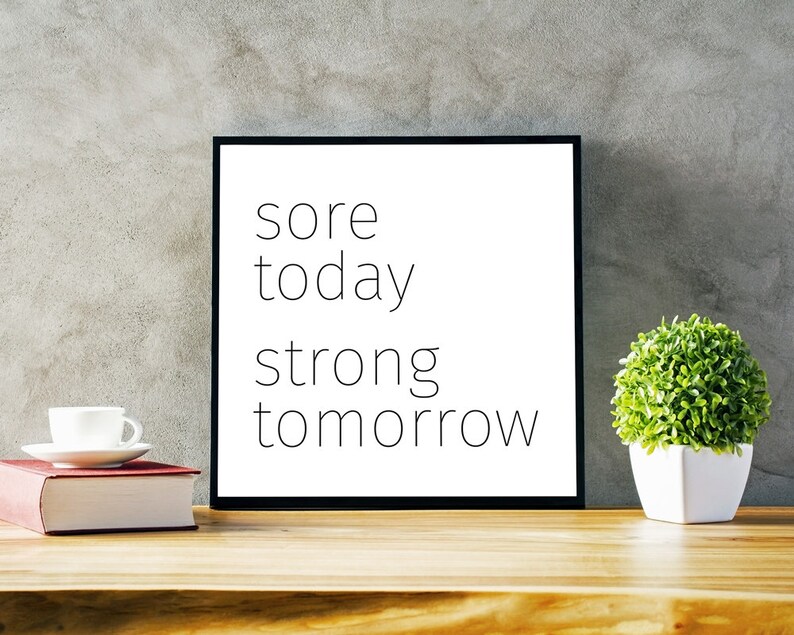 May include: A black framed motivational poster with the text "sore today strong tomorrow" on a white background. The poster is sitting on a wooden table with a white coffee cup and a potted plant.