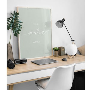May include: A framed print with a light green background and white text that reads "Your own words". The print is hanging on a white wall above a desk with a laptop, a lamp, and a plant.