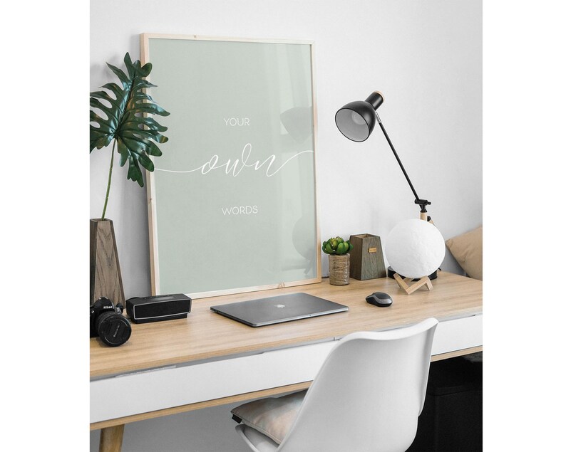May include: A framed print with a light green background and white text that reads "Your own words". The print is hanging on a white wall above a desk with a laptop, a lamp, and a plant.