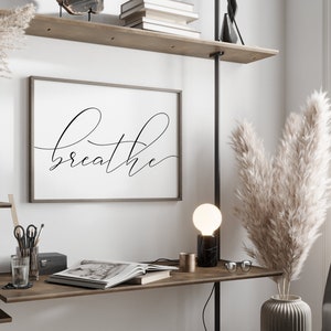 May include: A black and white framed print with the word "breathe" written in cursive script. The print is hanging on a white wall above a wooden desk with a lamp, a vase of dried pampas grass, and a book.
