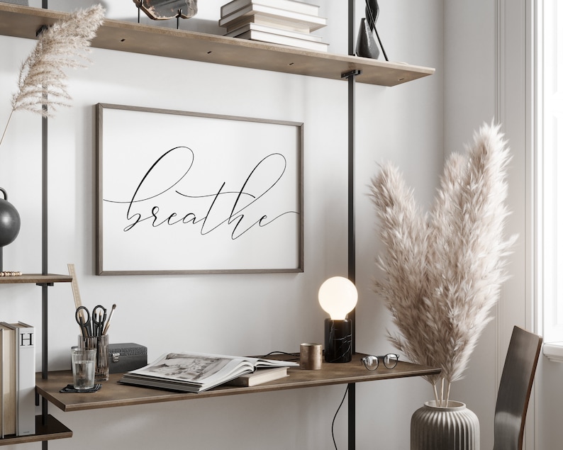 May include: A black and white framed print with the word "breathe" written in cursive script. The print is hanging on a white wall above a wooden desk with a lamp, a vase of dried pampas grass, and a book.