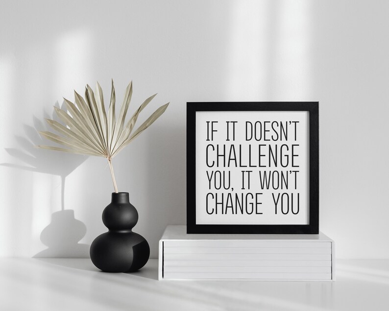 May include: Black framed motivational quote print with the text "If it doesn't challenge you, it won't change you." The print is sitting on a white shelf with a black vase and dried palm leaves.