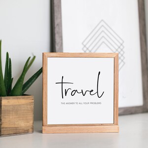 May include: A framed print with the word "Travel" in black script, with the phrase "the answer to all your problems" below it. The frame is light brown. A potted aloe plant and a framed geometric print are in the background.
