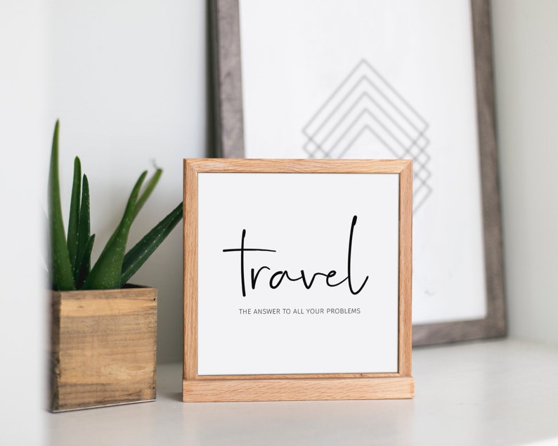 May include: A framed print with the word "Travel" in black script, with the phrase "the answer to all your problems" below it. The frame is light brown. A potted aloe plant and a framed geometric print are in the background.