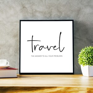 May include: A framed print with the word "travel" in a handwritten font, with the text "THE ANSWER TO ALL YOUR PROBLEMS" below. The frame is black, and the background is white. A small potted plant and a book are on the wooden surface.