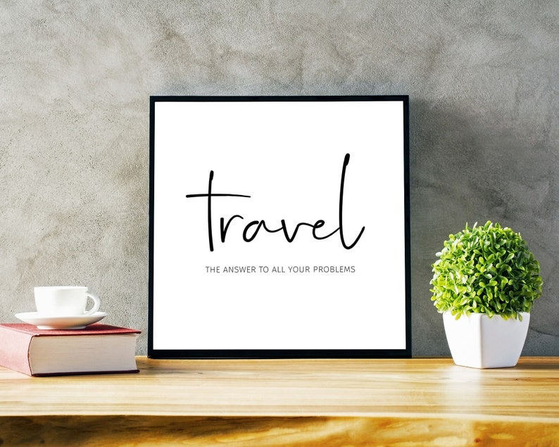 May include: A framed print with the word "travel" in a handwritten font, with the text "THE ANSWER TO ALL YOUR PROBLEMS" below. The frame is black, and the background is white. A small potted plant and a book are on the wooden surface.