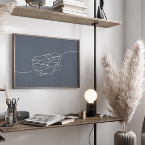 Puede incluir: Una moderna oficina en casa con una impresi&oacute;n enmarcada que presenta un dibujo lineal minimalista de libros apilados. La habitaci&oacute;n incluye un escritorio con una l&aacute;mpara, un libro abierto y objetos decorativos, con plantas altas y secas en un jarr&oacute;n.